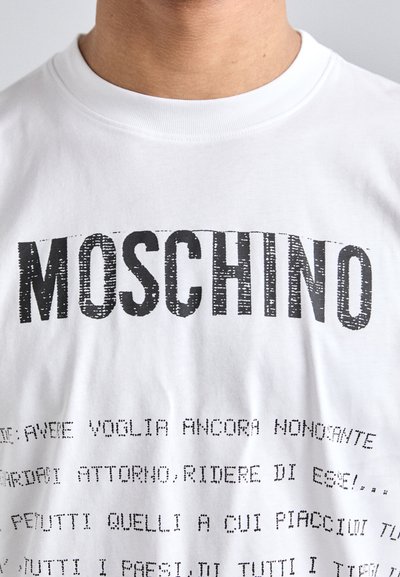 Hvit bomull T-skjorte med fet svart "MOSCHINO" tekst øverst og mindre svart tekst under i skrivemaskin skrifttype.