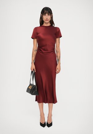 Woman with tattoos wearing a burgundy satin midi dress, black pointed heels, and holding a black handbag with gold detail against a white background.