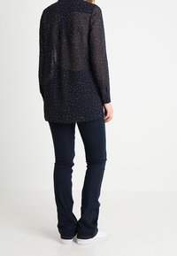 Mörknavy långärmad blus med vita prickar, semi-transparent tyg, ryggsöm och rundad nederkant, kombinerad med mörka jeans och vita skor.