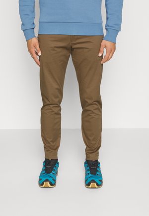 Mann trägt braune, schmal geschnittene Hose, blauen Pullover und blau-schwarze Sportschuhe, steht vor einem schlichten Hintergrund.