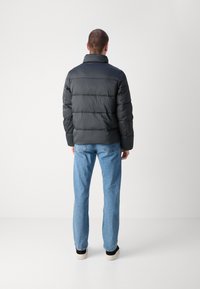 Giacca a vento nera con colletto alto, caratterizzata da sezioni trapuntate e una superficie liscia e lucida, indossata sopra jeans azzurri chiari con una vestibilità dritta.