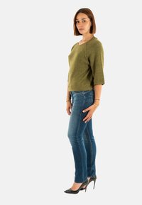 Pull vert olive à manches trois-quarts, associé à un jean slim bleu et des escarpins noirs. Tissu simple et texturé avec un minimum de détails.