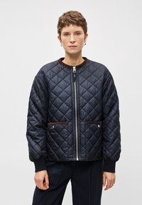 Melnā vītnē šūta bomber jaka ar brūniem akcentiem, rāvējslēdzēja priekšpuse un divas sānu kabatas. Iezīmējas ar ribotām aprocēm un apaļu kakla līniju. Gluda tekstūra.