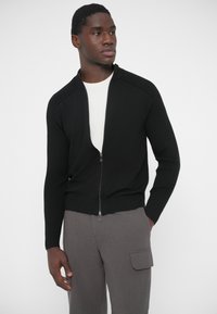 Čierny sveter na zips s rebrovanou textúrou, vysokým golierom a priliehavým strihom. Nosí sa cez biele tričko, v kombinácii so sivými nohavicami.