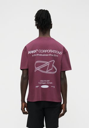 Άνδρας με dreadlocks φοράει μπορντό μπλουζάκι ARKK Corporations με λευκή γραμματοσειρά και λογότυπο, συνδυασμένο με μαύρο σορτς, γυρισμένος προς τα πίσω.