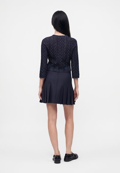 Femme aux longs cheveux noirs, debout de dos, portant un pull texturé foncé, une jupe plissée foncée et des mocassins noirs sur un fond blanc uni.