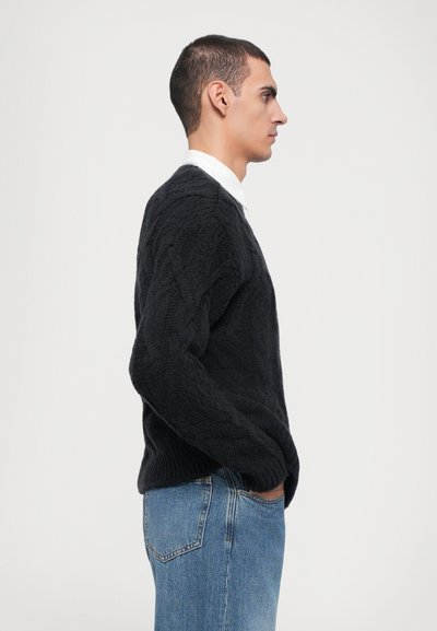 Black knitted sweater with cable pattern, round neck, and ribbed cuffs worn with light blue jeans. Side profile view. Smooth texture.