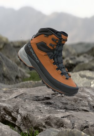 Botte de randonnée La Sportiva marron et noire avec étiquette Gore-Tex flottant au-dessus d'un terrain rocheux et d'un arrière-plan montagneux flou.