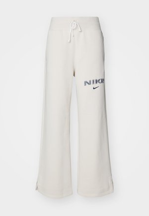 Pantalones de chándal anchos de color beige claro, con cinturilla elástica y cordón, aberturas laterales y logo bordado de "NIKE" en azul oscuro.