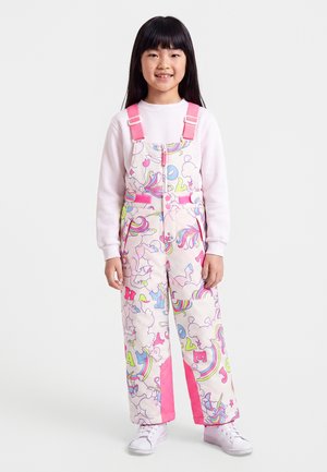 Meisje met lang donker haar draagt een witte trui en een roze patroon tuinbroek met eenhoorns, regenbogen en letters, staat en lacht.