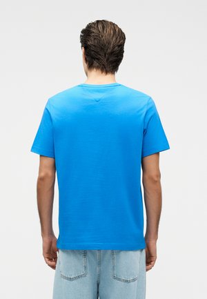 Mann mit kurzen braunen Haaren, der ein schlichtes blaues kurzärmliges T-Shirt und hellblaue Jeans trägt, und mit dem Rücken zu weißem Hintergrund steht.