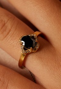 Gouden ring met een zwarte ovale steen, omringd door kleine heldere stenen. Glad band en textuur van de huid op de achtergrond.