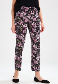 Femme portant un pantalon noir fuselé avec un motif floral rose et blanc, des ballerines noires et un haut sans manches noir.