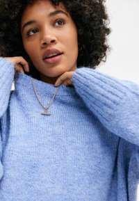 Hellblauer, gestrickter Pullover mit gerippter Struktur und weiten Ärmeln, kombiniert mit einer goldenen Kettenhalskette mit einem barförmigen Anhänger.