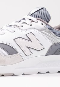 Närbild av en grå och vit New Balance-sneaker som visar strukturerad tyg, mocka, snören och "N"-logotypen på sidan.
