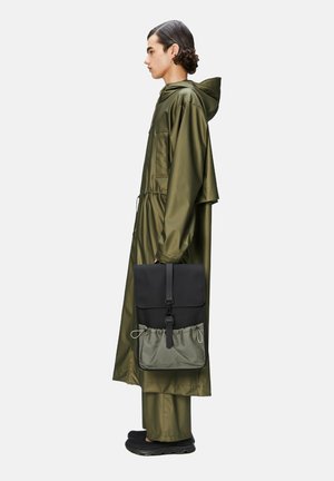 Chaqueta impermeable verde oliva con cordón, combinada con una mochila bicolor: sección superior negra y parte inferior verde con cierre de cordón.