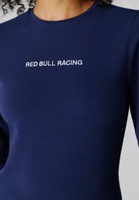 Temno modra dolga srajca z dolgimi rokavi, na prsih zgoraj z belim vezenjem napis RED BULL RACING, ki jo nosi oseba s kodrastimi lasmi.