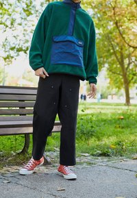 Grön fleecepullover med en blå dragkedjeficka, i kombination med svarta tapered byxor och röda sneakers. Texturerat tyg, avslappnad passform.