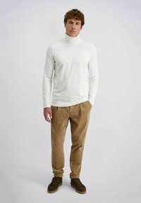 Man staat met zijn handen in zijn zakken, gekleed in een witte coltrui, bruine corduroy broek en donkerbruine sneakers tegen een effen lichte achtergrond.