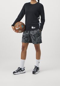 Chemise noire à manches longues, short noir à motifs, logo Nike blanc, tenant un ballon de basket. Baskets noires et blanches avec semelles texturées.