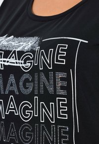 T-shirt nera con scollatura rotonda caratterizzata da testo bianco e strass argento che formano la parola "IMAGINE", creando un design grafico audace.