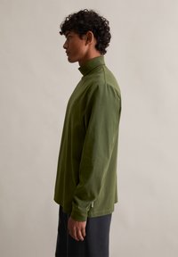 Maslinasto zelena majica s dugim rukavima i ovratnikom na dugme, izrađena od mekog materijala, s opuštenim krojem i bočnim prorezima na rubu za lakše kretanje.