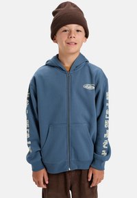 Jongen in een bruine gebreide beanie en een blauwe hoodie met ritssluiting en witte opdruk op de mouwen, staand tegen een effen lichte achtergrond.