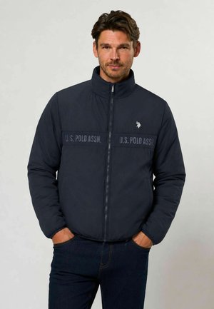 Homme portant une veste zippée bleu marine U.S. Polo Assn. avec les mains dans les poches, debout devant un fond uni clair.