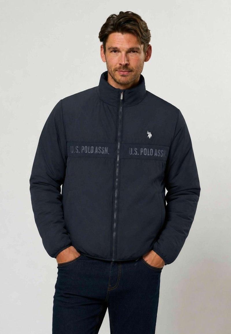 Homme portant une veste zippée bleu marine U.S. Polo Assn. avec les mains dans les poches, debout devant un fond uni clair.