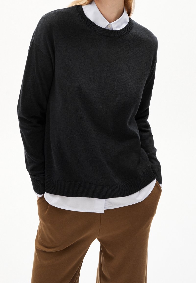 Pull-over noir à col rond avec poignets côtelés, superposé à une chemise blanche à col. Texture douce et coupe décontractée.