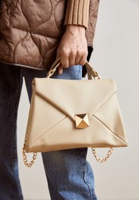 Beige læderhåndtaske med en struktureret form, der har en guld pyramide detalje, topbærerhåndtag og en aftagelig guld kædestrop.