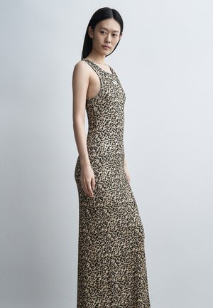 Kobieta z długimi czarnymi włosami, ubrana w bezrękawnikową, długą sukienkę w panterkę, stojąca bokiem na jednolitym jasnoszarym tle.