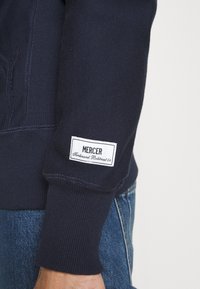 Sweatshirt bleu marine avec des poignets côtelés. Présente une étiquette blanche avec "MERCER" et une adresse. Texture lisse, tissu de poids moyen.