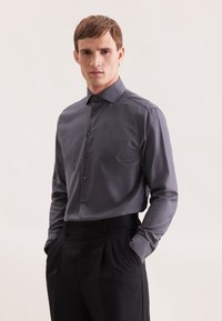 Camicia da uomo a maniche lunghe di colore blu navy con un piccolo motivo a quadretti bianchi, colletto a punta e tessuto morbido. Abbinata a pantaloni neri sartoriali.