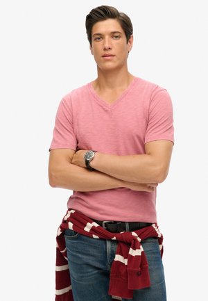 Camiseta rosa de escote en V, de mangas cortas; lleva un suéter de rayas rojas y blancas atado a la cintura; jeans azules; reloj plateado en la muñeca.