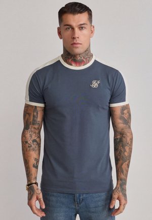 Mężczyzna z tatuażami na szyi i ramionach, ubrany w niebiesko-biały krótki t-shirt i niebieskie dżinsy, stojący na jednolitym tle.