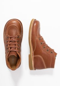 Bottes chelsea en cuir marron avec un upper texturé, bout rond, design à lacets, et des coutures blanches contrastantes avec une semelle en caoutchouc.