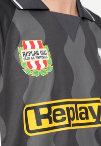 Γκρι φανέλα ποδοσφαίρου με κυματιστό μοτίβο, μαύρο γιακά, κίτρινο λογότυπο "Replay" και έμβλημα ασπίδας με κόκκινες και λευκές ρίγες.