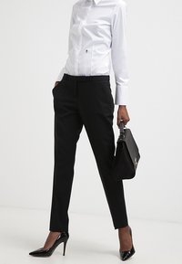 Pantalones negros ajustados con un corte slim, combinados con una camisa blanca de manga larga y tacones de aguja negros. Se sostiene un bolso estructurado negro.