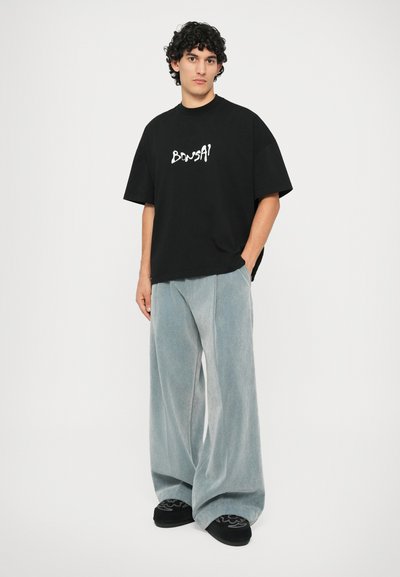 T-shirt noir oversize avec un texte graphique blanc, associé à un pantalon large bleu pâle et des chaussures noires arborant un design décoratif.