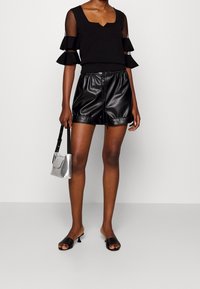 Schwarzes Oberteil mit transparenten, rüschierten Ärmeln, kombiniert mit hoch-taillierten schwarzen Shorts aus Kunstleder und schwarzen offenen Sandalen. Kleine silberne Handtasche.