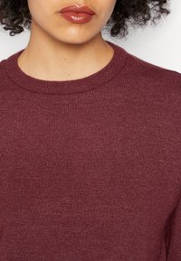 Kobieta z kręconymi włosami, ubrana w buraczkowy sweter z okrągłym dekoltem i ciemnoczerwoną szminką, pokazana od szyi do górnej części klatki piersiowej.