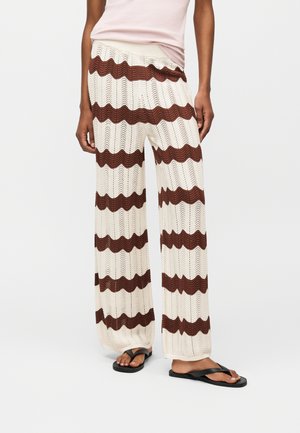 Άτομο που φορά πλεκτό παντελόνι σε εκρού και καφέ σε φαρδιά γραμμή με οριζόντιες κυματιστές ρίγες, συνδυασμένο με μαύρες σαγιονάρες και ροζ ανοιχτό τοπ.