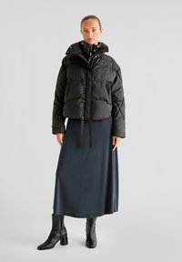 Sort puffy jakke med cropped design, blank overflade og trykknaplukning, parret med en lang mørkeblå nederdel og sorte hælede støvler.
