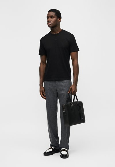 Άνδρας όρθιος, φοράει μαύρο μπλουζάκι, γκρι παντελόνι, μαύρα και άσπρα μοκασίνια και κρατά ένα μαύρο δερμάτινο χαρτοφύλακα.