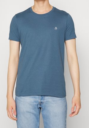 Person iført en blå t-shirt med rund hals og et lille hvidt logo på venstre bryst samt lyseblå jeans, står med afslappede arme.
