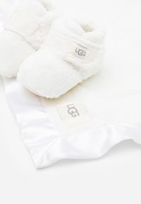 Białe, puszyste botki wykonane z sztucznego futra, z logo UGG, w zestawie z gładkim satynowym kocem z małą metką UGG.