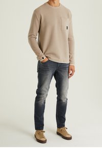 Muž ve béžové texturované košili s dlouhým rukávem, vybledlých modrých džínách a světlých teniskách, stojící s jednou rukou v kapse.