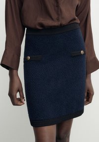 Ullkjol i marinblått med texturerad finish, med svarta detaljer och två framfickor prydda med metallknappar. Slimmad design, knälång.