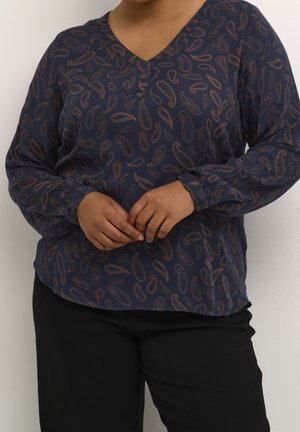 Personne portant un chemisier bleu marine à manches longues avec des motifs cachemire marron et un pantalon noir, les mains légèrement jointes devant.